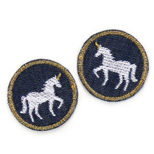 May include: Two white embroidered unicorn patches on a dark blue denim background with a gold thread outline.