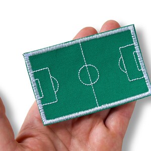 Könnte beinhalten: Ein rechteckiger, bestickter Aufnäher mit einem grünen Fußballfeld-Design, bei dem weiße Sticklinien die Spielfeldbegrenzungen, den Mittelkreis und die Torräume darstellen, eingefasst von einem weißen Stickrand um den gesamten Umfang.
