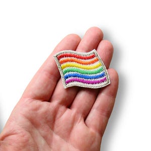 Può includere: Una piccola toppa ricamata rettangolare caratterizzata da un design arcobaleno ondulato e multicolore con strisce orizzontali rosse, arancioni, gialle, verdi, blu e viola, rifinita con un bordo cucito grigio chiaro su tutti e quattro i lati.