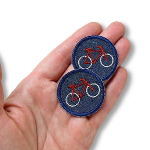 May include: Two round denim patches with a red bicycle embroidered on each. The bicycle has white wheels and a red frame. The patches have a white border and are held in a hand.