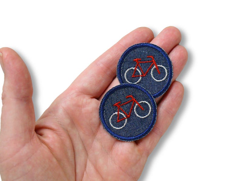 May include: Two round denim patches with a red bicycle embroidered on each. The bicycle has white wheels and a red frame. The patches have a white border and are held in a hand.
