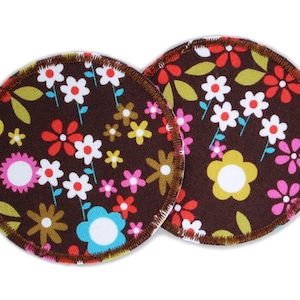 Könnte beinhalten: Zwei braune Still-Einlagen aus Stoff mit einem Blumenmuster. Das Muster zeigt weiße, gelbe, rote, rosa und blaue Blumen auf braunem Hintergrund.