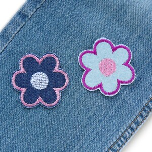 May include: Two fabric flower patches sewn onto a blue denim background. One flower is blue with a white center and pink edges. The other flower is pink with a white center and blue edges.