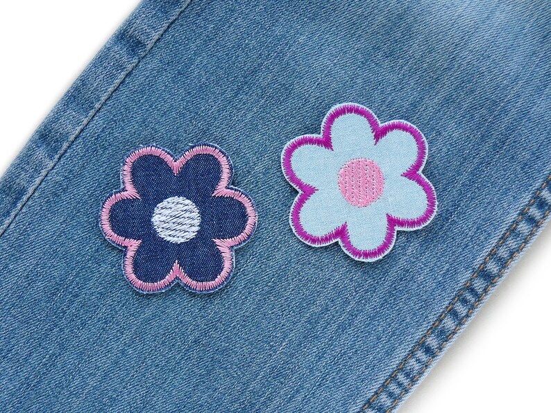 May include: Two fabric flower patches sewn onto a blue denim background. One flower is blue with a white center and pink edges. The other flower is pink with a white center and blue edges.