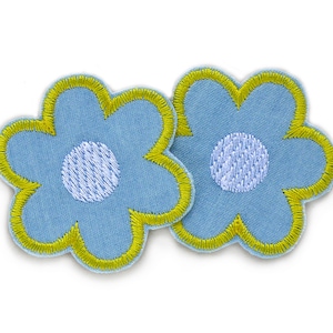 Könnte beinhalten: Zwei blaue Stoffblumen-Patches mit gelben Umrandungen und weißen Zentren. Die Blumen haben einen Retro-Stil.