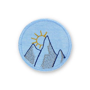 May include: A circular, light blue embroidered patch depicting a mountain range and a yellow sun. The mountains are grey with blue outlines, and the sun has yellow rays. The patch has a stitched border, suitable for jackets, bags, or other fabric items.