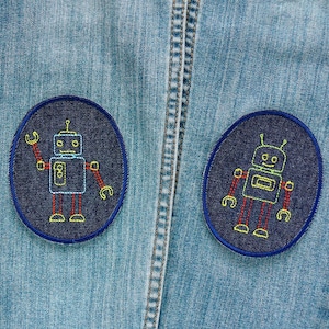 Könnte beinhalten: Zwei aufgestickte Roboter-Patches auf hellblauem Denim. Jeder Patch zeigt ein Roboter-Design mit roten, gelben und blauen Nähten. Die Patches sind oval mit einem dunkelblauen Rand, ideal zum Dekorieren von Kleidung oder Accessoires.