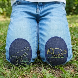 May include: Light blue denim jeans with dark blue knee patches. Each patch features a green and yellow embroidered dinosaur design. The jeans are worn by a person sitting on green grass.