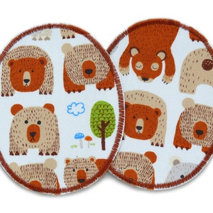 Op de afbeelding: Twee ronde stoffen patches met een bruine gestikte rand. Elke patch heeft een herhalend patroon van cartoonberen in verschillende poses en kleuren, waaronder bruin, beige en wit, tegen een lichtblauwe achtergrond. Een boom, wolken en paddenstoelen zijn ook inbegrepen.