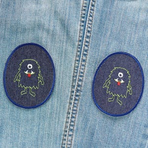 Könnte beinhalten: Hellblaue Jeans mit zwei ovalen Aufnähern. Jeder Aufnäher zeigt ein marineblaues Monsterdesign mit einem einzelnen Auge, grünen Umrissen und einer roten Zunge. Die Aufnäher sind blau umrandet.