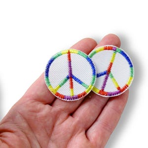 Könnte beinhalten: Zwei runde, weiße, gestickte Aufnäher mit dem Peace-Zeichen. Das Symbol ist mit Regenbogenfarben gestickt: rot, orange, gelb, grün, blau und lila. Die Aufnäher werden in einer Hand vor weißem Hintergrund gehalten.