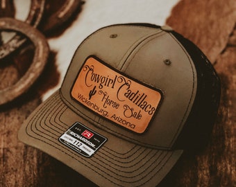 May include: A green and black trucker hat with a brown leather patch that says "Cowgirl Cadillacs Horse Sale Wickenburg, Arizona". The hat has a Richardson 112 label.