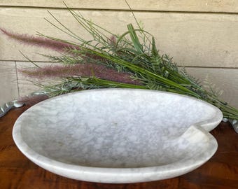 May include: A white marble bowl with a unique, asymmetrical shape. The bowl has a smooth, polished surface and a slightly irregular edge.