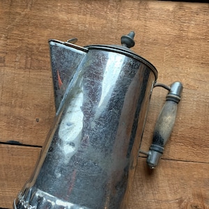 May include: A vintage silver metal pitcher with a wooden handle. The pitcher has a decorative embossed pattern around the bottom.