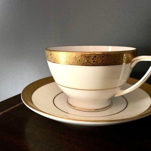 May include: A white teacup and saucer with a gold trim and intricate design. The cup and saucer are on a dark brown surface.