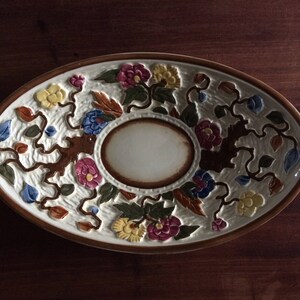 May include: An oval ceramic serving dish with a white background and a floral design. The design features brown vines, green leaves, and colorful flowers in shades of pink, yellow, blue, and brown. The center of the dish has a smaller, circular, white serving area.
