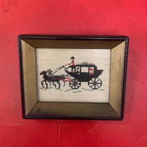 May include: A framed cross-stitch embroidery depicting a black horse-drawn carriage with a driver in a red coat. The carriage is detailed with white and red accents and is set against a cream-colored background.
