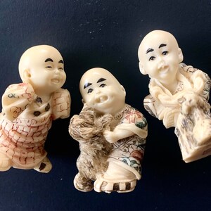 May include: Three small ivory figurines of smiling monks, each wearing traditional robes with intricate patterns. The monk on the left is holding a small animal, the monk in the center is holding a small animal, and the monk on the right is holding a scroll.