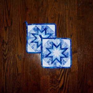 May include: Two square quilted potholders with a starburst design in shades of blue and white. Each potholder has a loop for hanging. The potholders are approximately 8 inches square.