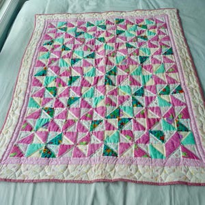 May include: A handmade quilt with a patchwork design. The quilt features a repeating pattern of triangles in shades of pink, teal, and floral prints, bordered by a pink and cream trim. The quilt is likely for a baby or child.