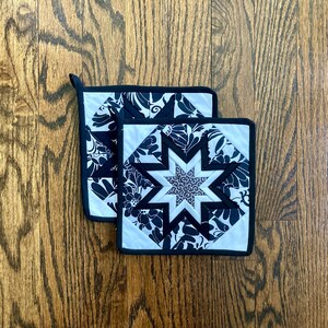 May include: Two black and white quilted pot holders with a star design. The pot holders are made of fabric with a floral pattern.