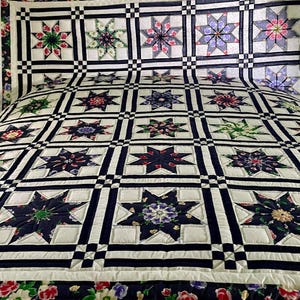 May include: A bed quilt featuring a repeating star pattern within a grid of dark navy blue and white fabric strips. Each star block contains a unique floral print in shades of red, green, purple, and blue, bordered by solid white and navy fabric.