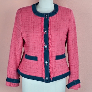 May include: A pink tweed jacket with a blue denim trim and fringe detail. The jacket has a button closure and two pockets.
