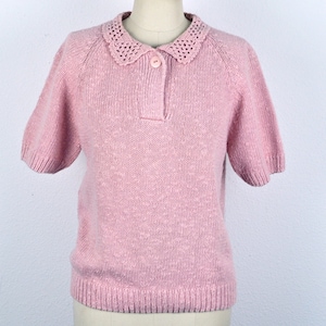 May include: A pink knitted sweater with a short sleeve and a crocheted collar. The sweater has a button closure at the neck.