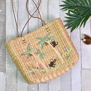 May include: Woven straw tote bag with brown leather handles. The bag features embroidered palm trees, monkeys, and flowers. The bag is rectangular and has a brown leather strap.