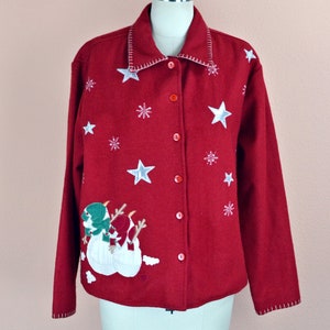 Puede incluir: Cárdigan rojo con botones con estrellas y copos de nieve blancos bordados en la parte delantera. El cárdigan presenta un diseño de muñeco de nieve con sombreros verdes y bufandas rojas.