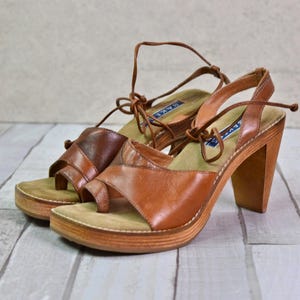 May include: A pair of brown leather sandals with wooden heels. The sandals have a platform sole and straps that wrap around the ankle. The heels are approximately 3 inches high.