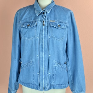 Puede incluir: Una chaqueta vaquera azul claro con cierre de cremallera y varios bolsillos. La chaqueta tiene un cuello abotonado y una etiqueta de marca en el pecho.