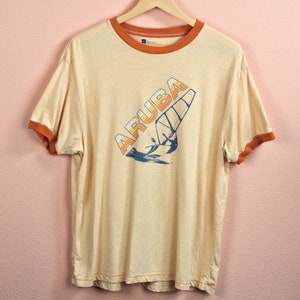 May include: A light yellow t-shirt with an orange and blue graphic of a sailboat and the word "Aruba" on the front. The shirt has a contrasting orange ringer.