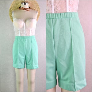 May include: Mint green high-waisted shorts with a pleated front. The shorts are displayed on a mannequin with a white lace bustier and a straw hat. The shorts have an elastic waistband and a tailored fit, perfect for a vintage or retro look.