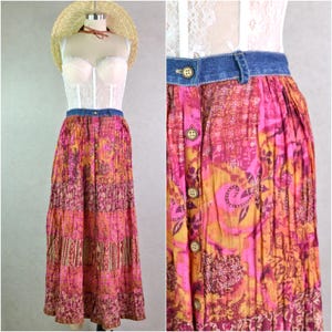 May include: A long, tiered skirt with a denim waistband and wooden buttons. The skirt features a vibrant floral pattern in shades of pink, orange, and brown. The skirt is displayed on a mannequin.