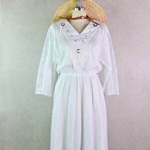 May include: White dress with long sleeves, a V-neck, and fringe detailing. The dress has a fitted waist and a flowing skirt. A straw hat is placed on top of the mannequin head. The dress is displayed on a mannequin.