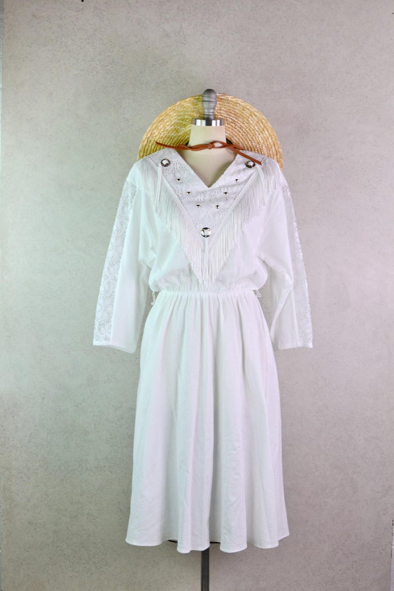 May include: White dress with long sleeves, a V-neck, and fringe detailing. The dress has a fitted waist and a flowing skirt. A straw hat is placed on top of the mannequin head. The dress is displayed on a mannequin.