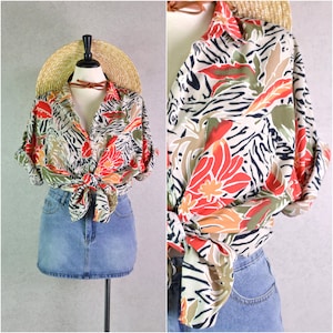 Könnte beinhalten: Ein Hemd mit Knöpfen und einem lebendigen Blumen- und Zebrastreifenmuster in Rot, Orange, Grün und Beige. Das Hemd ist an der Taille gebunden und wird mit einem Jeansrock kombiniert. Ein Strohhut und ein braunes Halsband ergänzen den Look.