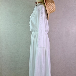 May include: A white long-sleeve dress with lace detailing on the sleeves and bodice, displayed on a mannequin. A straw hat with a brown band rests on the mannequin's head, adding a rustic touch. The dress has an elastic waist and a flowing skirt.