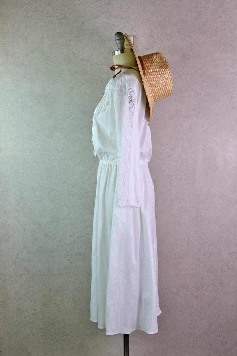 May include: A white long-sleeve dress with lace detailing on the sleeves and bodice, displayed on a mannequin. A straw hat with a brown band rests on the mannequin's head, adding a rustic touch. The dress has an elastic waist and a flowing skirt.
