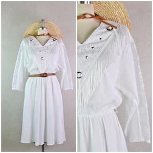May include: White vintage dress with long sleeves, a V-neck, and a brown belt. The dress features lace detailing on the sleeves and a fringed yoke with decorative metal accents. A straw hat is placed behind the dress form.