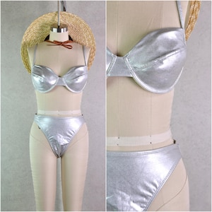 May include: A silver metallic bikini set displayed on a mannequin. The two-piece swimsuit includes a structured bra top and high-waisted bottoms. A straw hat with a brown ribbon is placed on top of the mannequin's head.