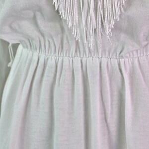 May include: Close-up of a white garment with a gathered waist and a fringed neckline. The fringe is made of long, thin strands of white material. The fabric appears to be a lightweight, possibly cotton or linen, with soft folds.