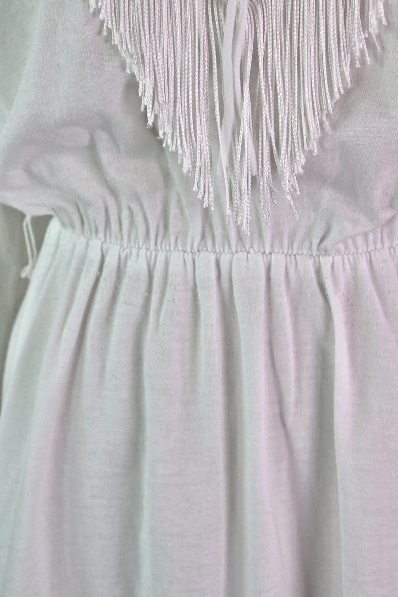 May include: Close-up of a white garment with a gathered waist and a fringed neckline. The fringe is made of long, thin strands of white material. The fabric appears to be a lightweight, possibly cotton or linen, with soft folds.