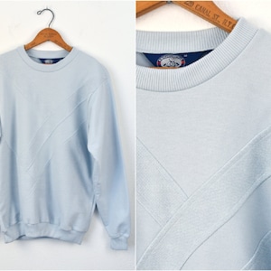 Peut inclure: Un sweat-shirt bleu clair à col rond et manches longues. Le devant présente un motif diagonal texturé. Le sweat-shirt est suspendu à un cintre en bois. L'étiquette du col indique "Progressive".