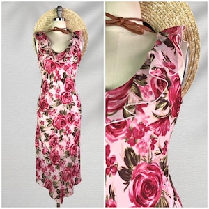 May include: A sleeveless, floral print dress with a ruffled neckline and a fitted silhouette. The dress features a pattern of large, red roses on a light pink background. A straw hat and brown leather neckband are also visible.