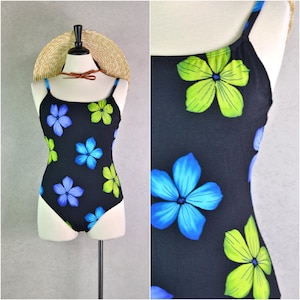 May include: Black one-piece swimsuit with a floral pattern. The swimsuit features blue and green flowers on a black background. The swimsuit has thin straps and a scoop neckline. A straw hat with a brown leather band is on top.