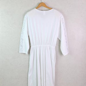 May include: A white dress with long sleeves and a cinched waist hangs on a wooden hanger. The sleeves feature lace detailing on the lower arms. The dress is made of a soft, flowing fabric and is displayed against a light gray wall.