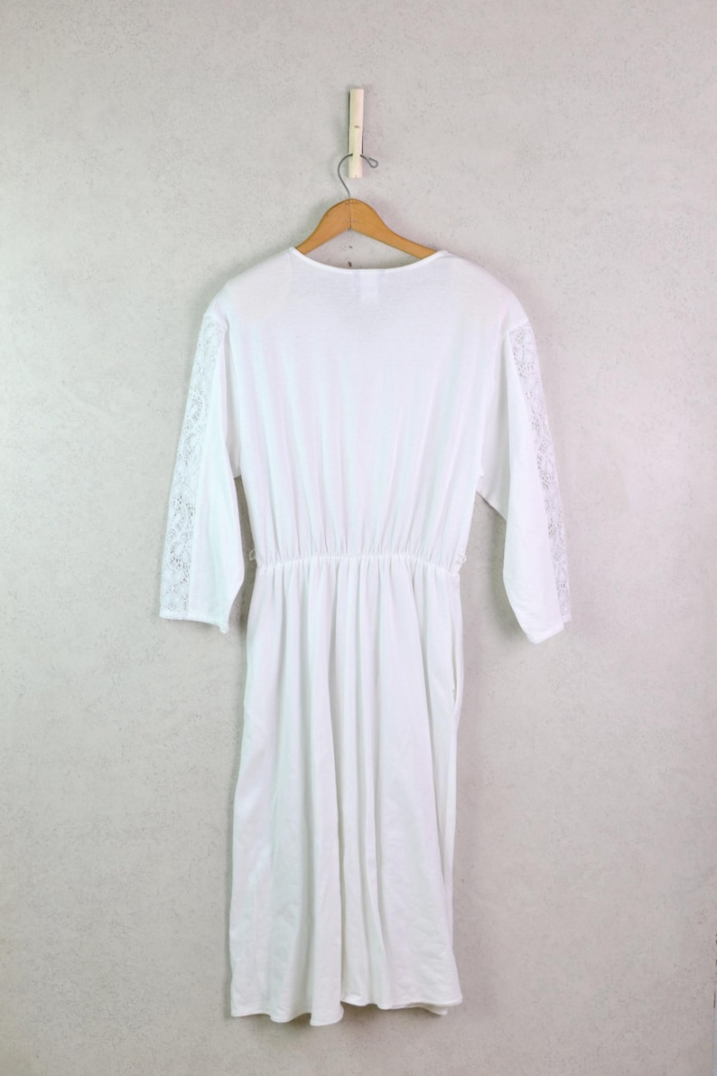 May include: A white dress with long sleeves and a cinched waist hangs on a wooden hanger. The sleeves feature lace detailing on the lower arms. The dress is made of a soft, flowing fabric and is displayed against a light gray wall.