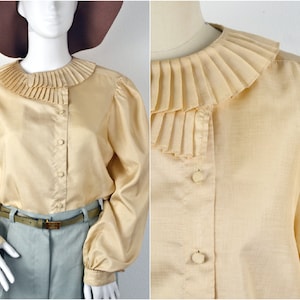 May include: A cream coloured blouse with a large, pleated, ruffled collar. The blouse has long sleeves with gathered cuffs and a button-up front.
